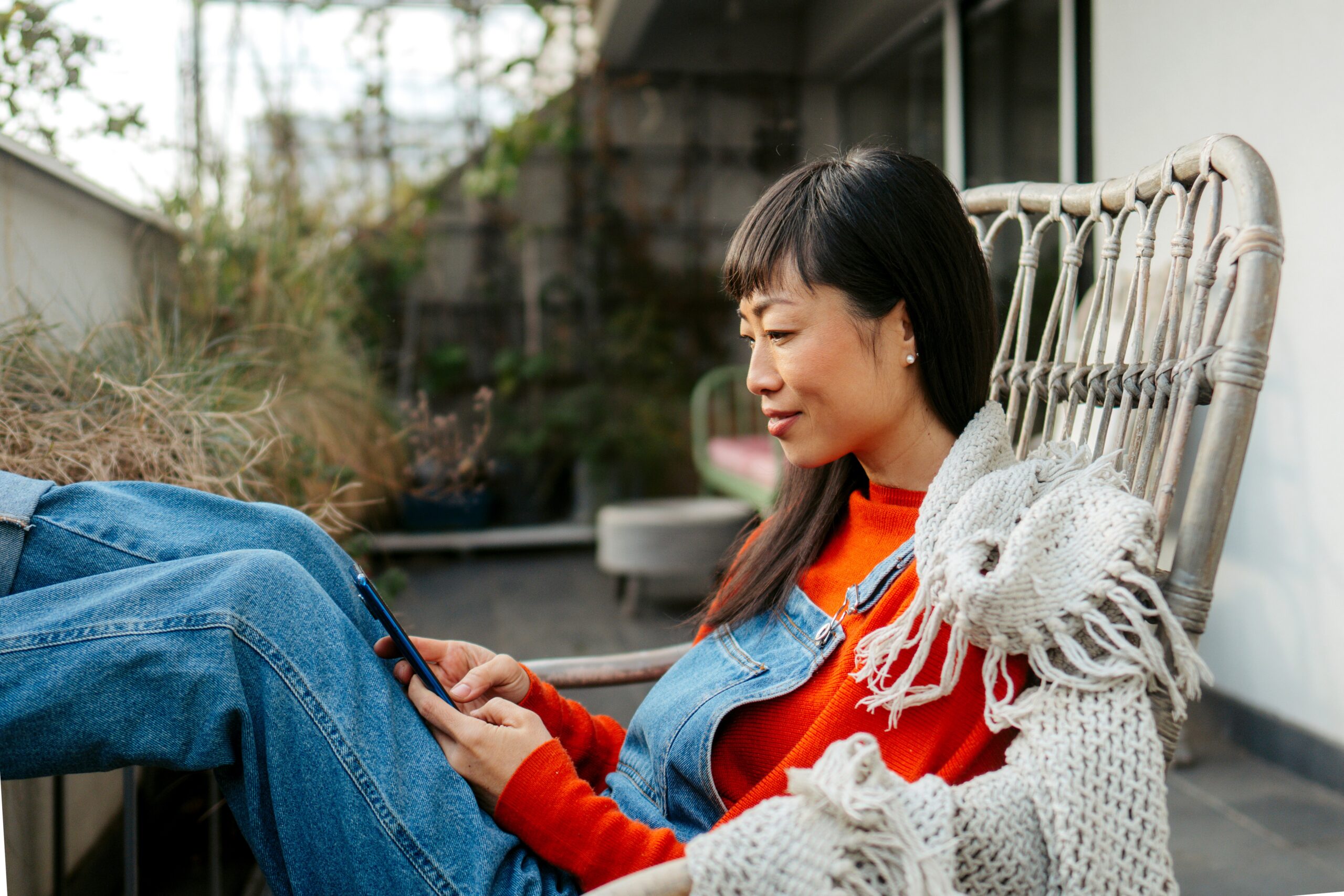 Young adult woman sitting outside on patio chair while looking at phone with smile on face